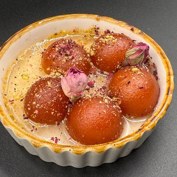 Five Gulab Jamun, an Indian dessert, in a white bowl with syrup, rose petals, and nuts.