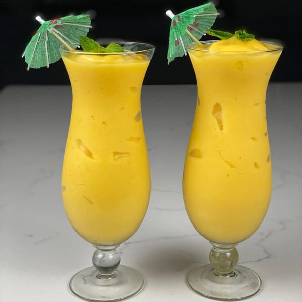 Two tall glasses of bright yellow mango lassi, garnished with mint and small green umbrellas.